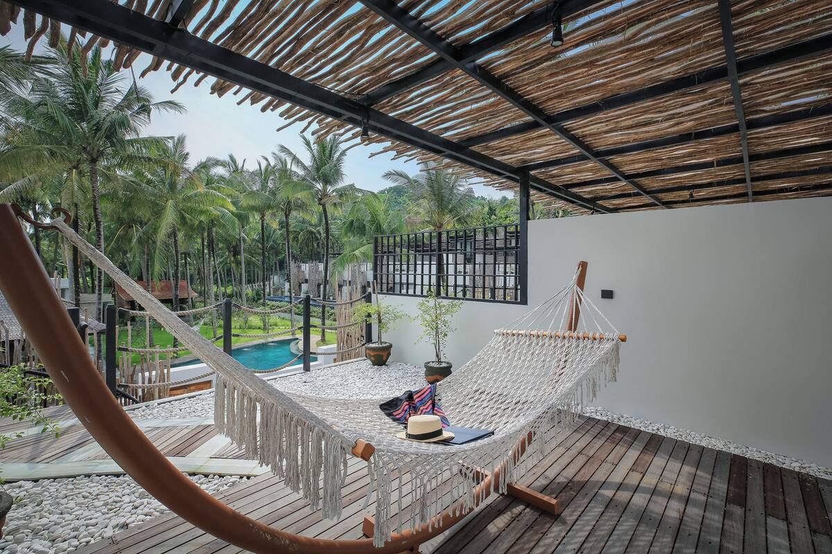 A hammock on a wooden deck with a view of palm trees and a pool.