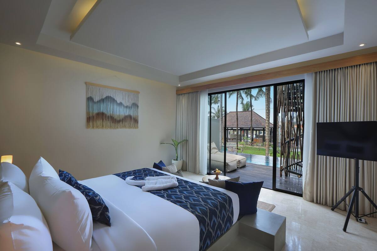 A modern hotel room with a large bed, a TV, and a balcony with a view of the pool and palm trees.