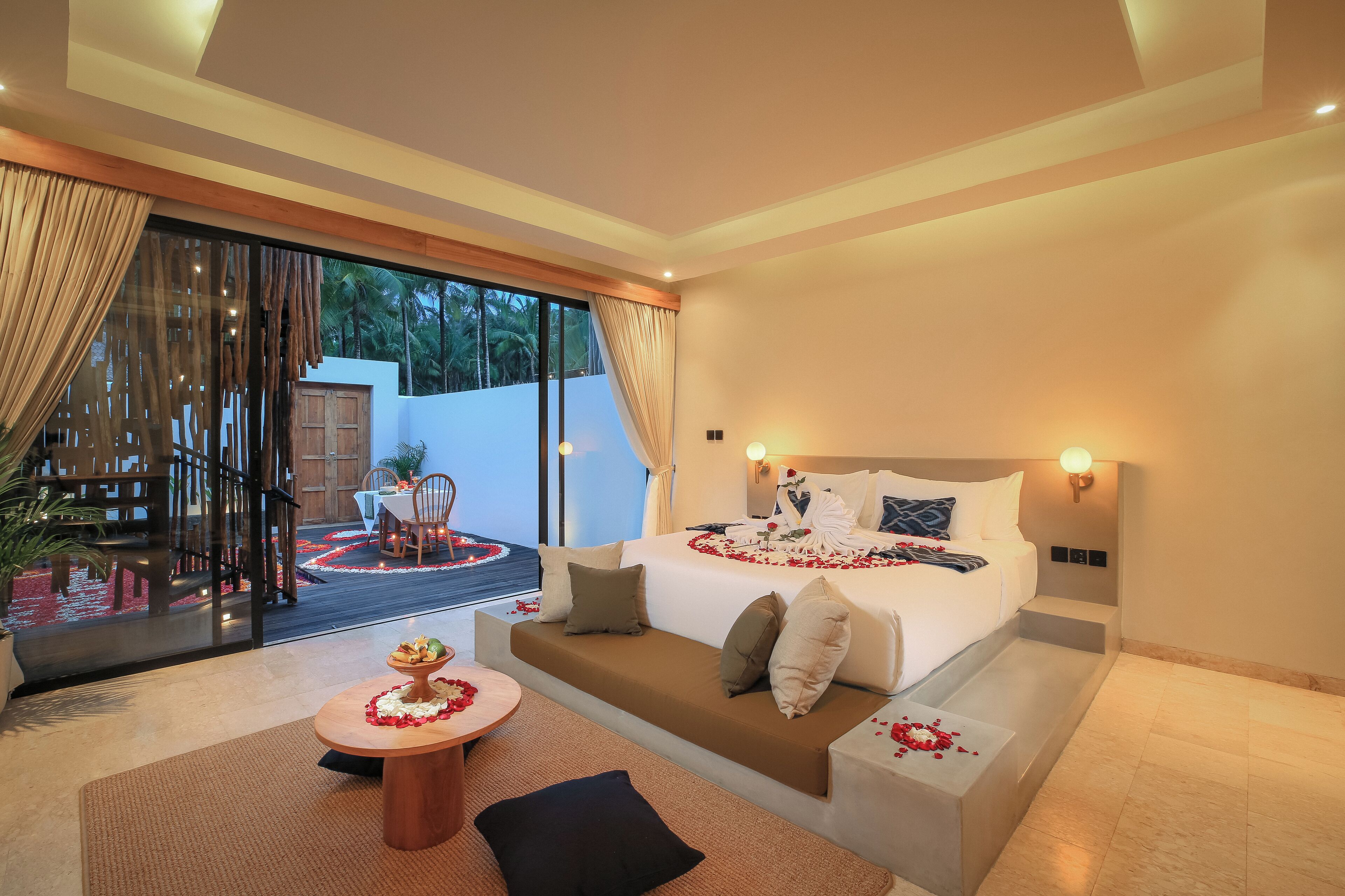 A modern bedroom with a large bed, a sofa, a small table with a fruit bowl, and a view of a garden through a sliding door.