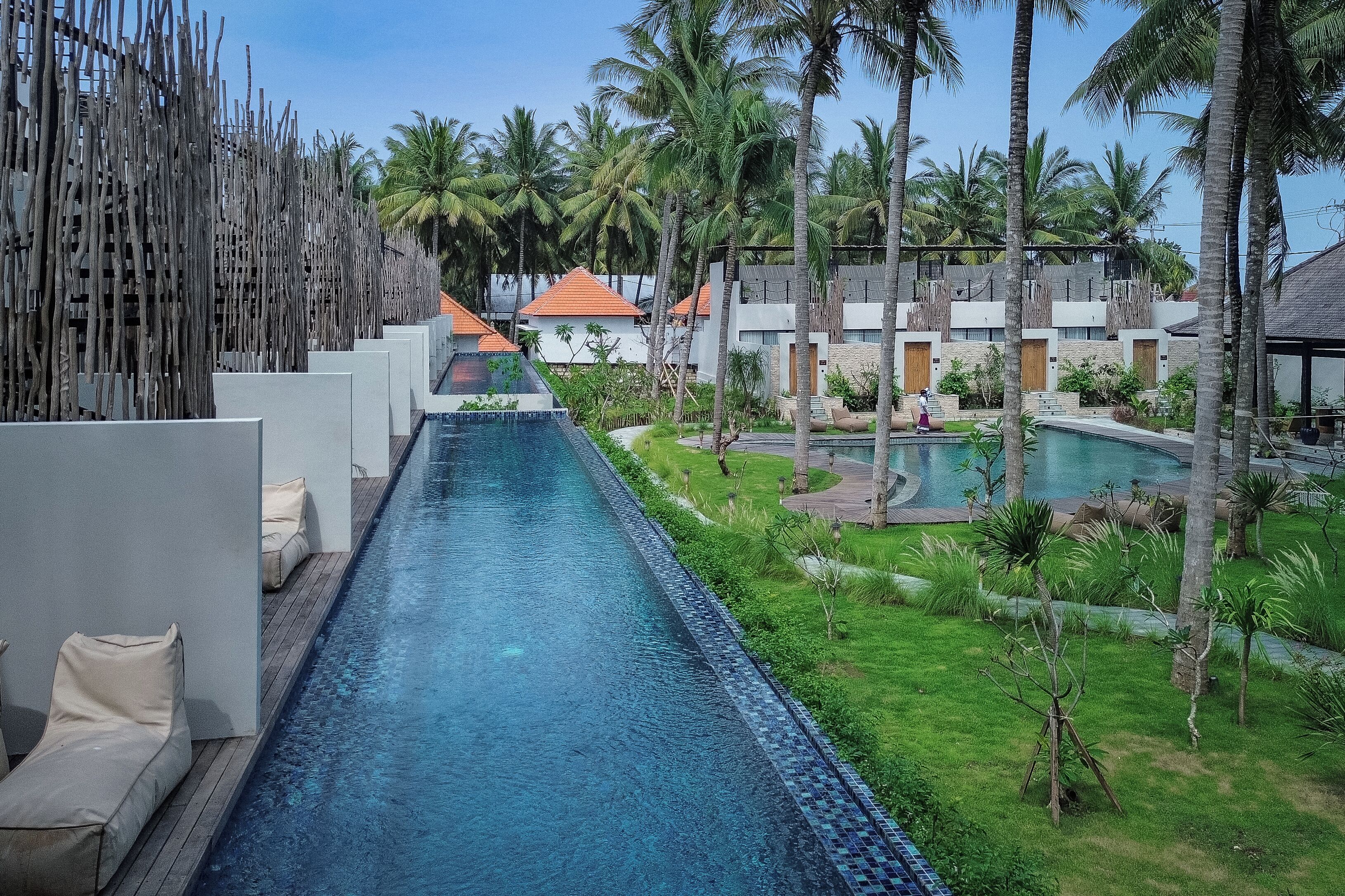 Una piscina llarga amb gandules, envoltada d'arbres de palm i un edifici modern amb teulada d'estil clàssic.