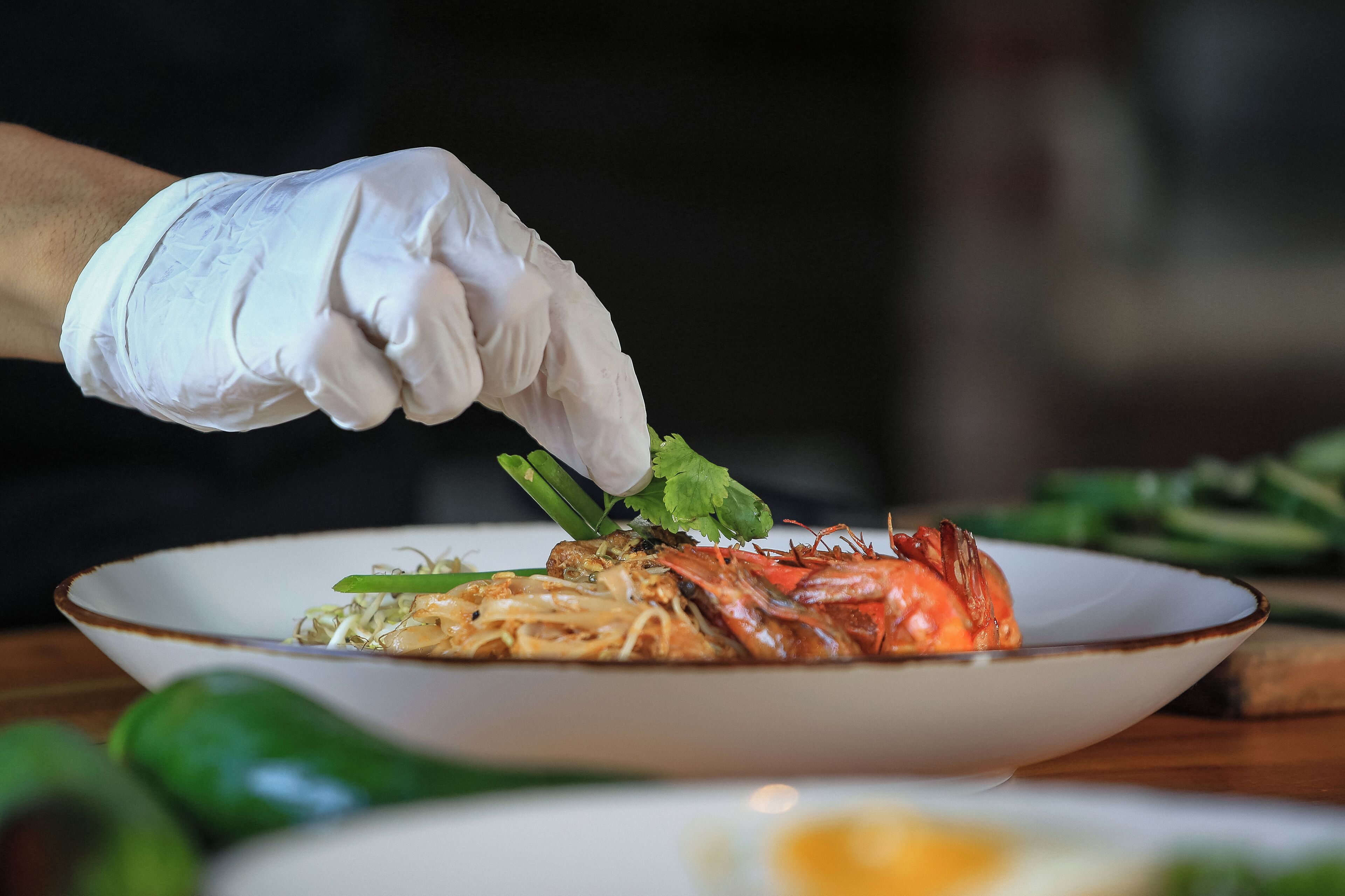 A person wearing a white glove placing a green vegetable on a plate with noodles and a cooked crab.