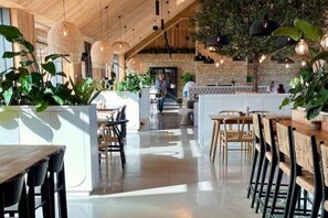 A spacious indoor area with wooden tables and chairs, a central seating area with a white cushioned bench, and hanging pendant lights. - Large barn with hot-tub in the English countryside

 (Oakham)