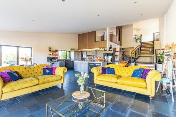 A spacious living area with two yellow sofas, a glass coffee table, and a kitchen in the background. - Large barn with hot-tub in the English countryside

 (Oakham)