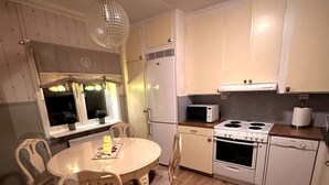 Een compacte keuken met witte kasten, een magnetron, een fornuis en een spoelbak. Er is een eettafel met stoelen en een kroonluchter.