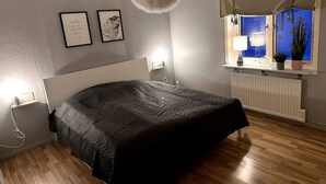 Een slaapkamer met een bed, nachtlampjes, een radiator, een raam met uitzicht en twee ingelijste foto's aan de muur.