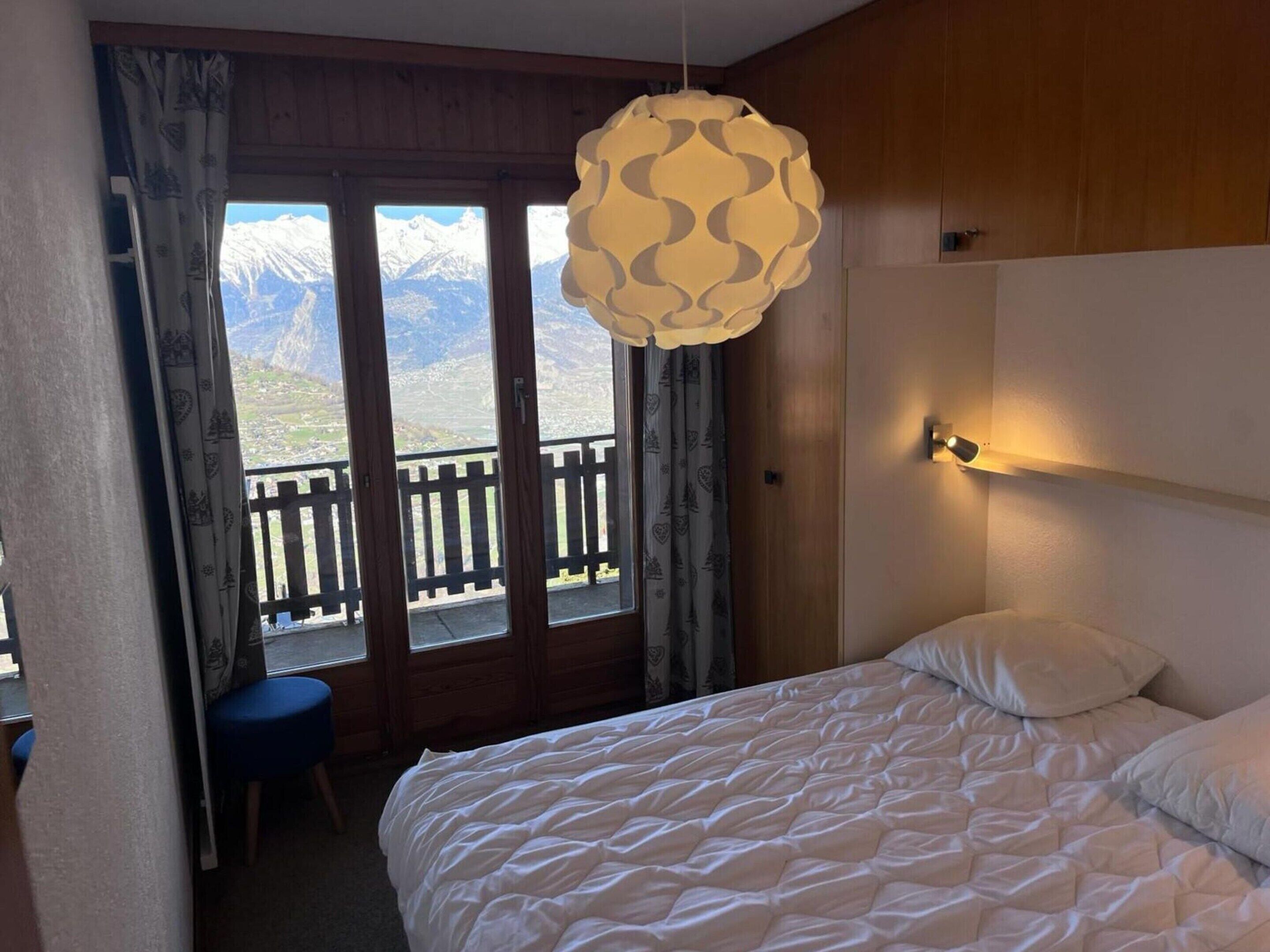 A bedroom with a bed, a hanging light fixture, a view of mountains through a window, and a wooden floor.