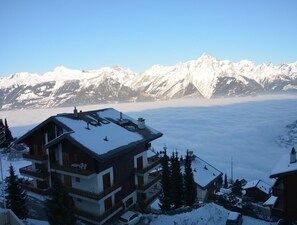 Ein mehrstöckiges Gebäude mit verschneitem Dach, umgeben von verschneiten Bäumen und Bergen im Hintergrund.