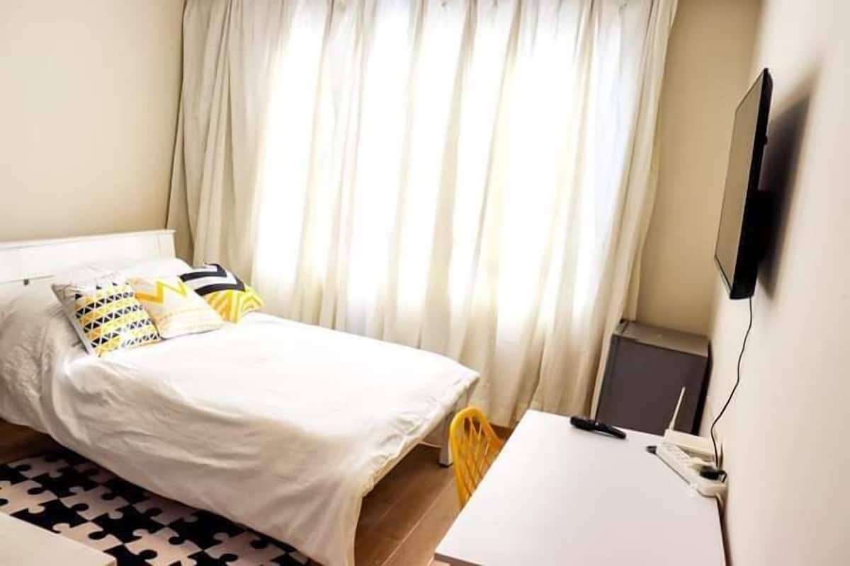 A bedroom with a bed, a desk, a chair, a TV, and a window with curtains.