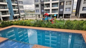 A swimming pool with a tiled deck, surrounded by a fence and greenery. In the background, there are multi-story buildings with balconies.