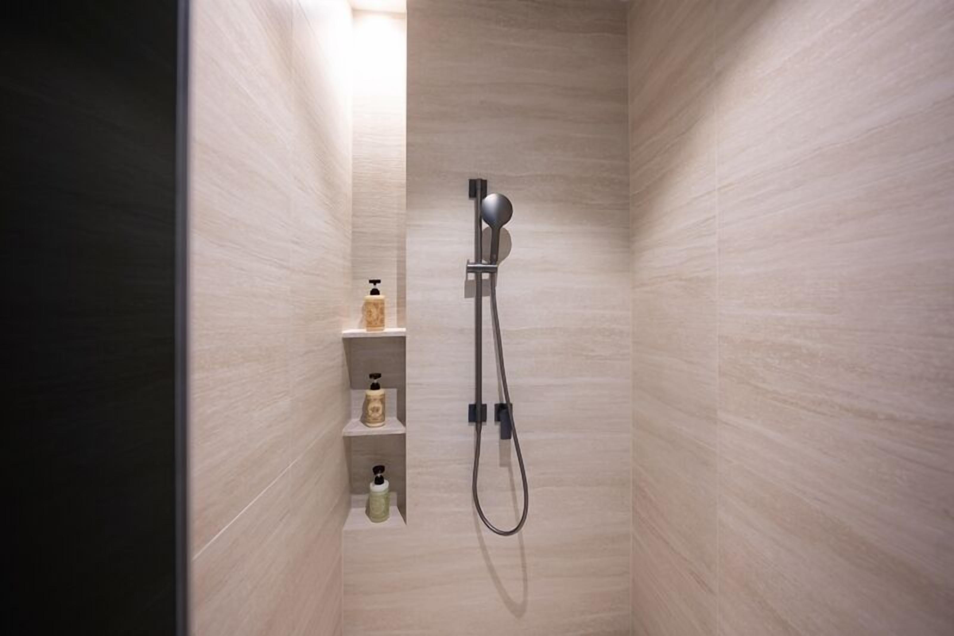 Un espace douche étroit doté d’un pommeau de douche à main et d’un robinet mural. On y trouve trois étagères contenant des articles de toilette.