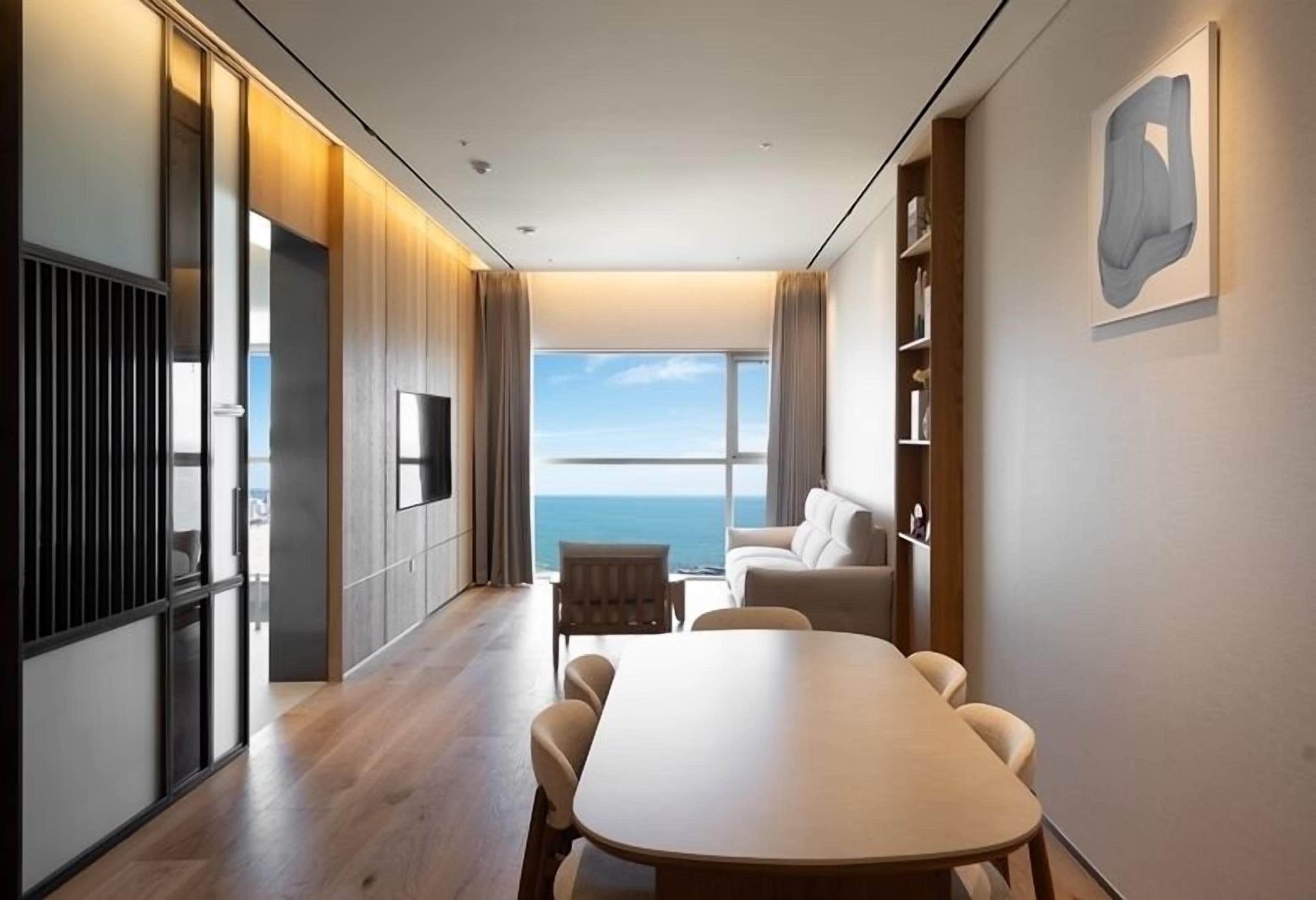 A modern hotel room with a large window offering a view of the sea, a dining area with a table and chairs, and a seating area with a sofa.