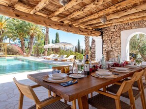 An outdoor dining area with a pool, wooden pergola, stone walls, and palm trees. - Impressive Farmhouse With Pool (Santa Eulalia del Rio)