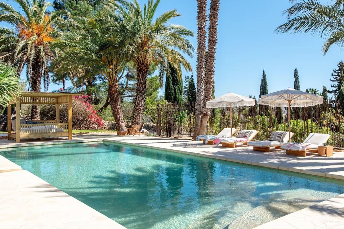 A clear swimming pool surrounded by lounge chairs and umbrellas, with palm trees and a wooden pergola in the background.