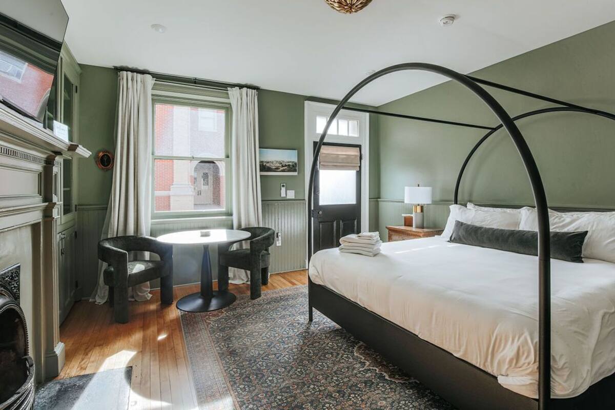 A bedroom with a four-poster bed, a round table with two chairs, a fireplace, and a large window with curtains.