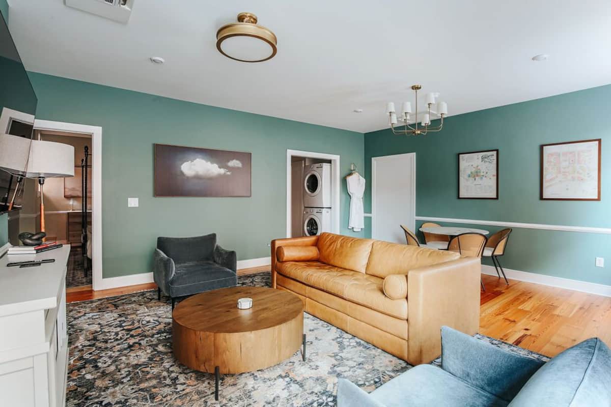 A living room with a beige sofa, a coffee table, and a gray armchair. There is a washing machine in the background.