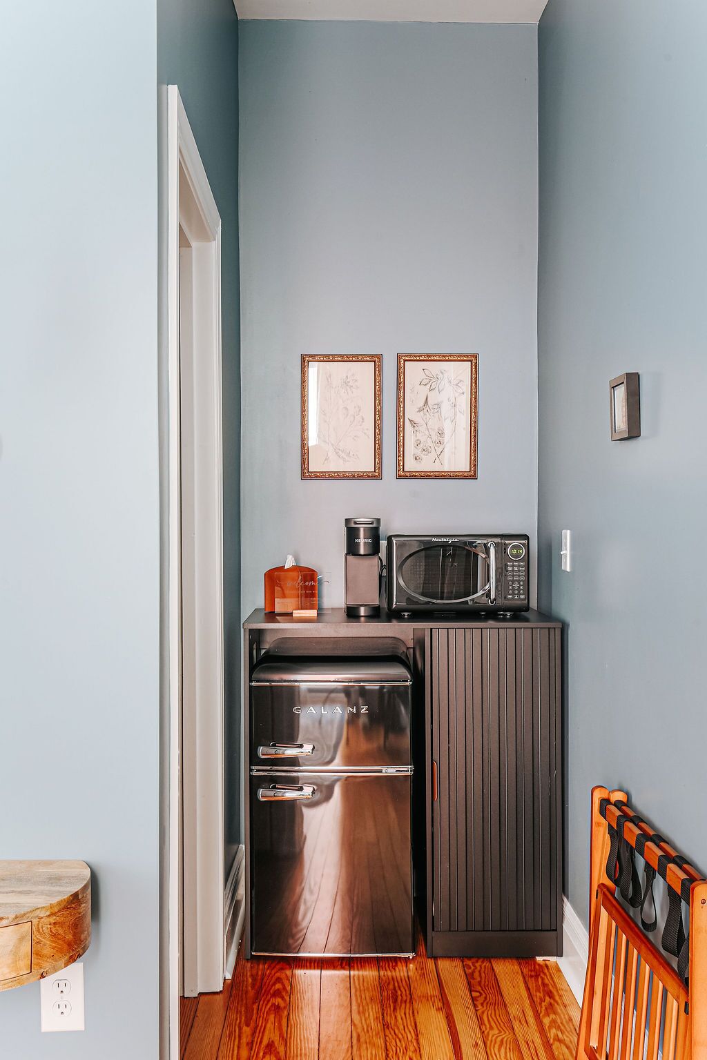 Une cuisine compacte équipée d’une cuisinière intégrée, d’un four à micro-ondes et d’une machine à café. Deux tableaux encadrés ornent le mur, ainsi qu’un petit meuble et un plancher en bois.