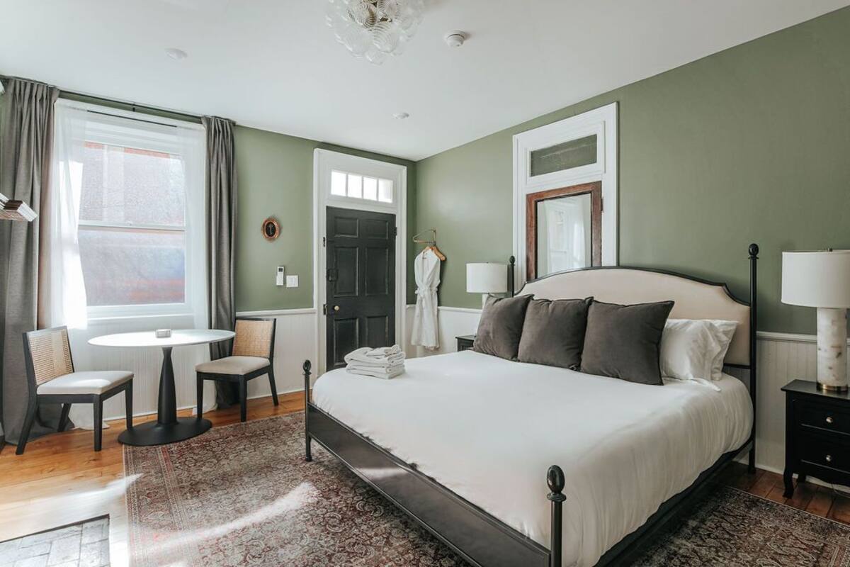 A bedroom with a large bed, a small table, two chairs, a window with curtains, and a door.