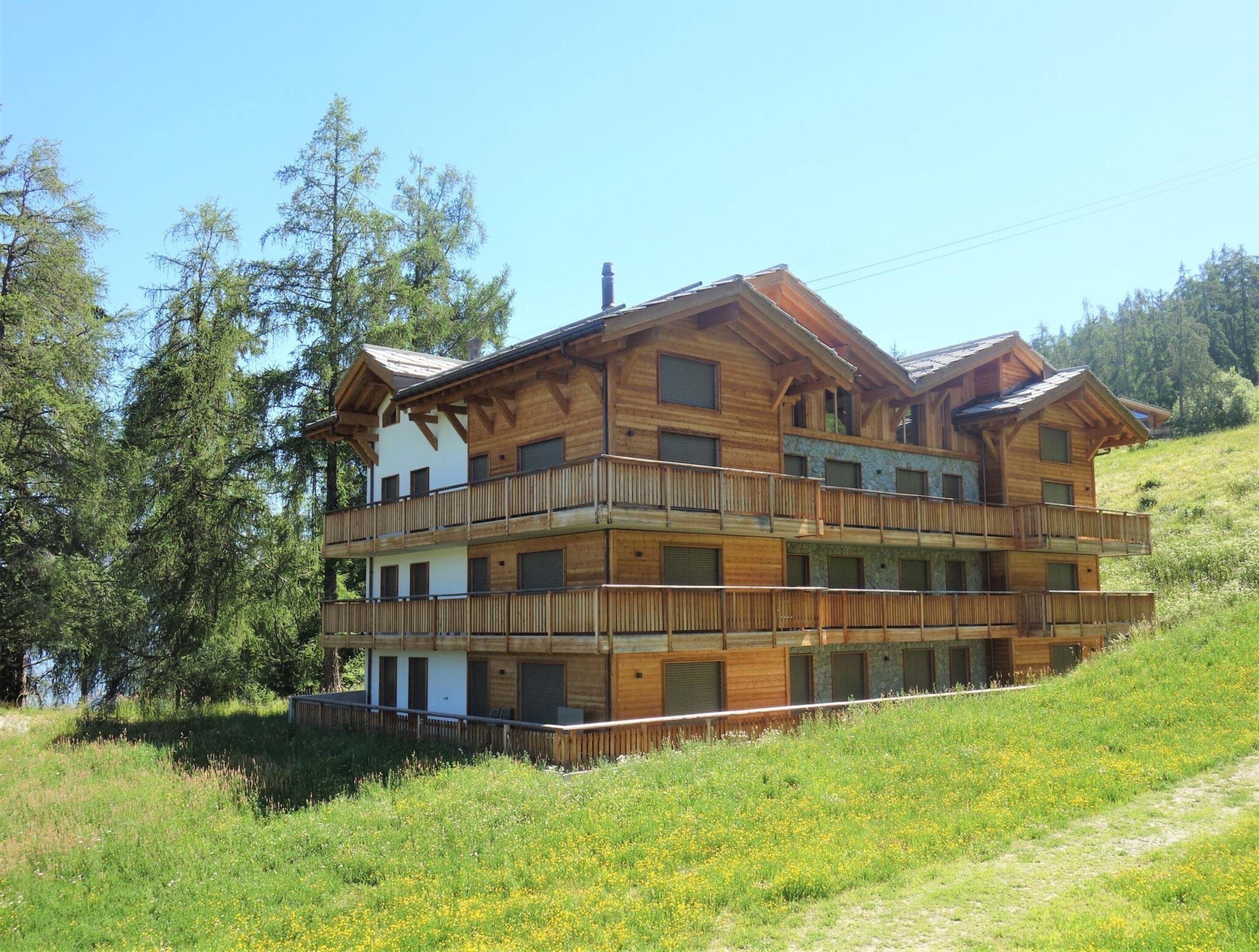 Un chalet en bois à plusieurs étages, doté de balcons, situé sur une colline et offrant une vue sur les arbres et un plan d’eau.