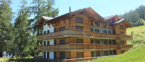 Un chalet en bois à plusieurs étages, doté de balcons, situé sur une colline et offrant une vue sur les arbres et un plan d’eau.