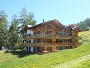 A multi-story wooden chalet with balconies, situated on a hillside with a view of trees and a body of water. - Luxury Apartment With View (Veysonnaz)