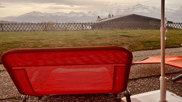 Une chaise rouge sous un parasol en chaume, une clôture en métal et, au loin, une vue sur la montagne.