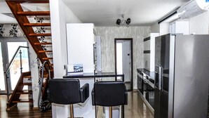 A modern kitchen with a stainless steel refrigerator, a built-in oven, and a microwave. There are two black bar stools and a small island with a sink. A wooden staircase with a floral wallpaper design leads to another room. - Holiday Apartment in Nature (Frenstat pod Radhostem)