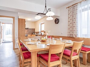 A dining area with a wooden table set for six, red cushioned chairs, a chandelier, and a kitchen in the background.