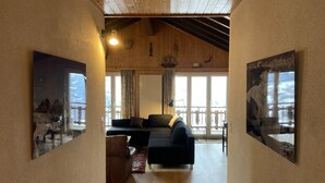 A spacious living area with a high ceiling, wooden beams, a sofa, a small table, and a view through a glass door to the outside. - Peaceful Apartment With Amenities (Veysonnaz)