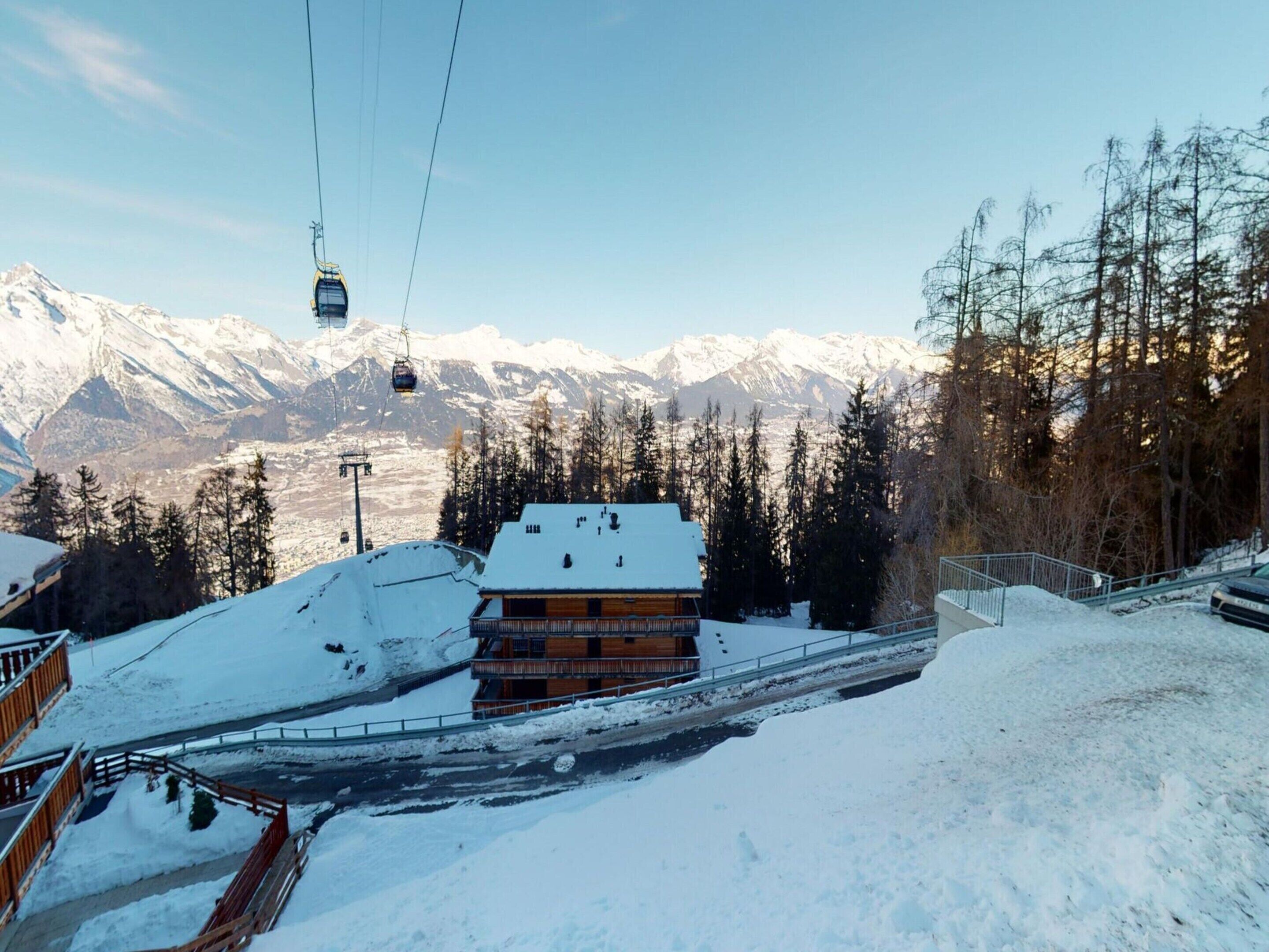 A snow-covered mountain landscape with a ski lift, a wooden building with a snow-covered roof, and a forested area.