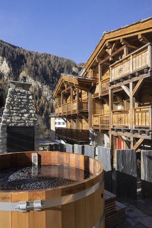 Una cabaña de madera con chimenea de piedra, un jacuzzi y balcones de madera con vistas a una zona montañosa.