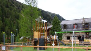 A playground with climbing structures and a bench, surrounded by a fence, with a mountain and a building in the background.
