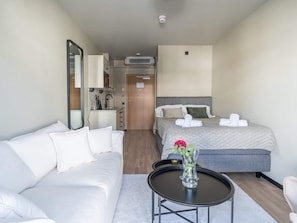 A hotel room with a bed, a sofa, a small table with a vase, a mirror, a kitchenette, and a door leading to another room.