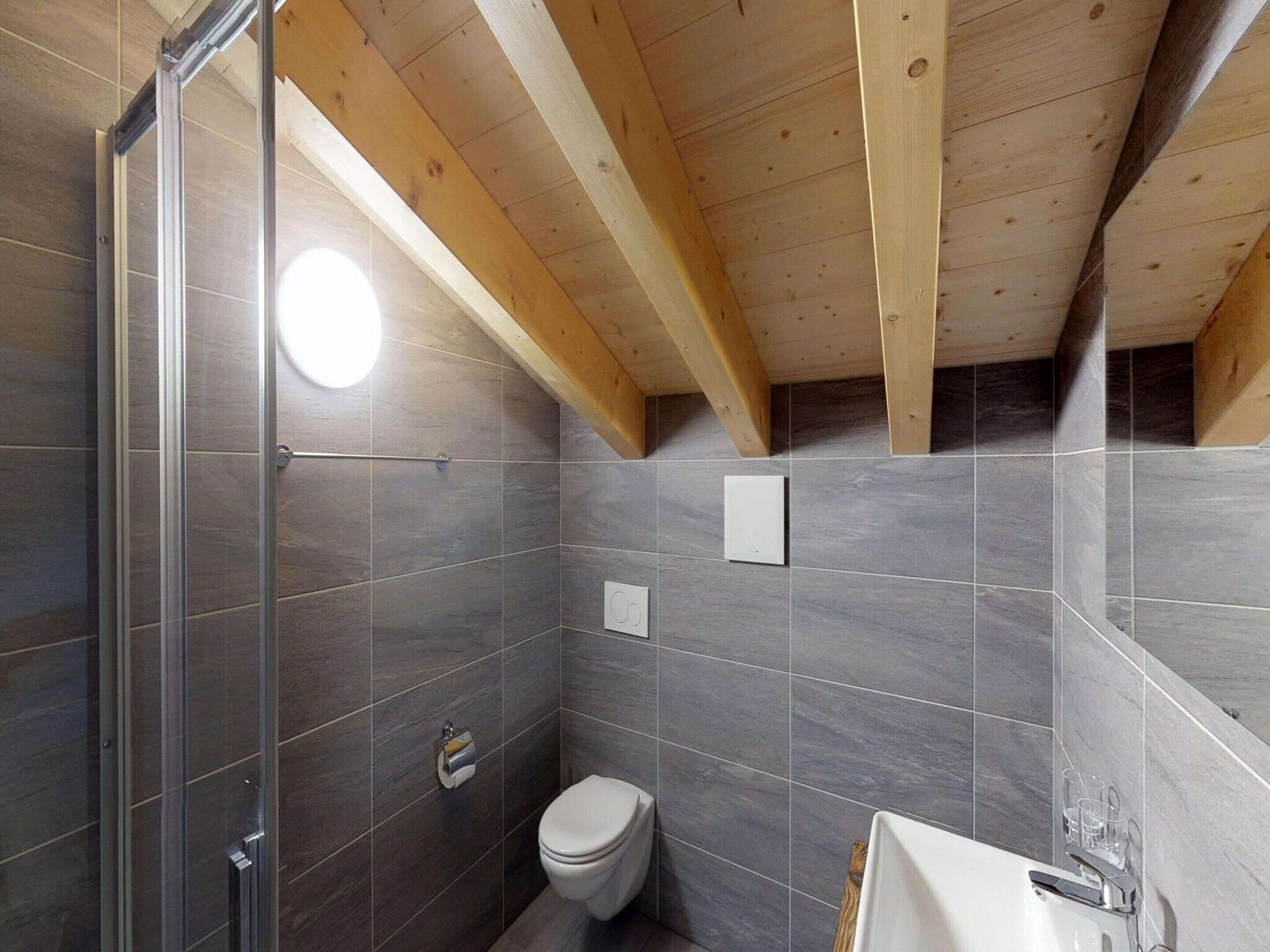 A bathroom with grey tiles, a toilet, and a shower with glass doors.