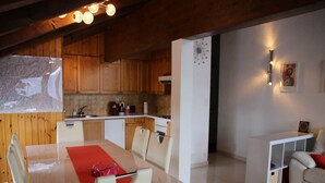 Una cocina moderna con armarios de madera, un comedor con mesa y sillas, y un mueble blanco pequeño con cojín rojo.