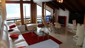 Una sala de estar moderna con un sofá blanco, cojines rojos, una mesa de centro de vidrio y una alfombra roja. Hay un ventanal con vistas al cielo y un televisor en la pared.