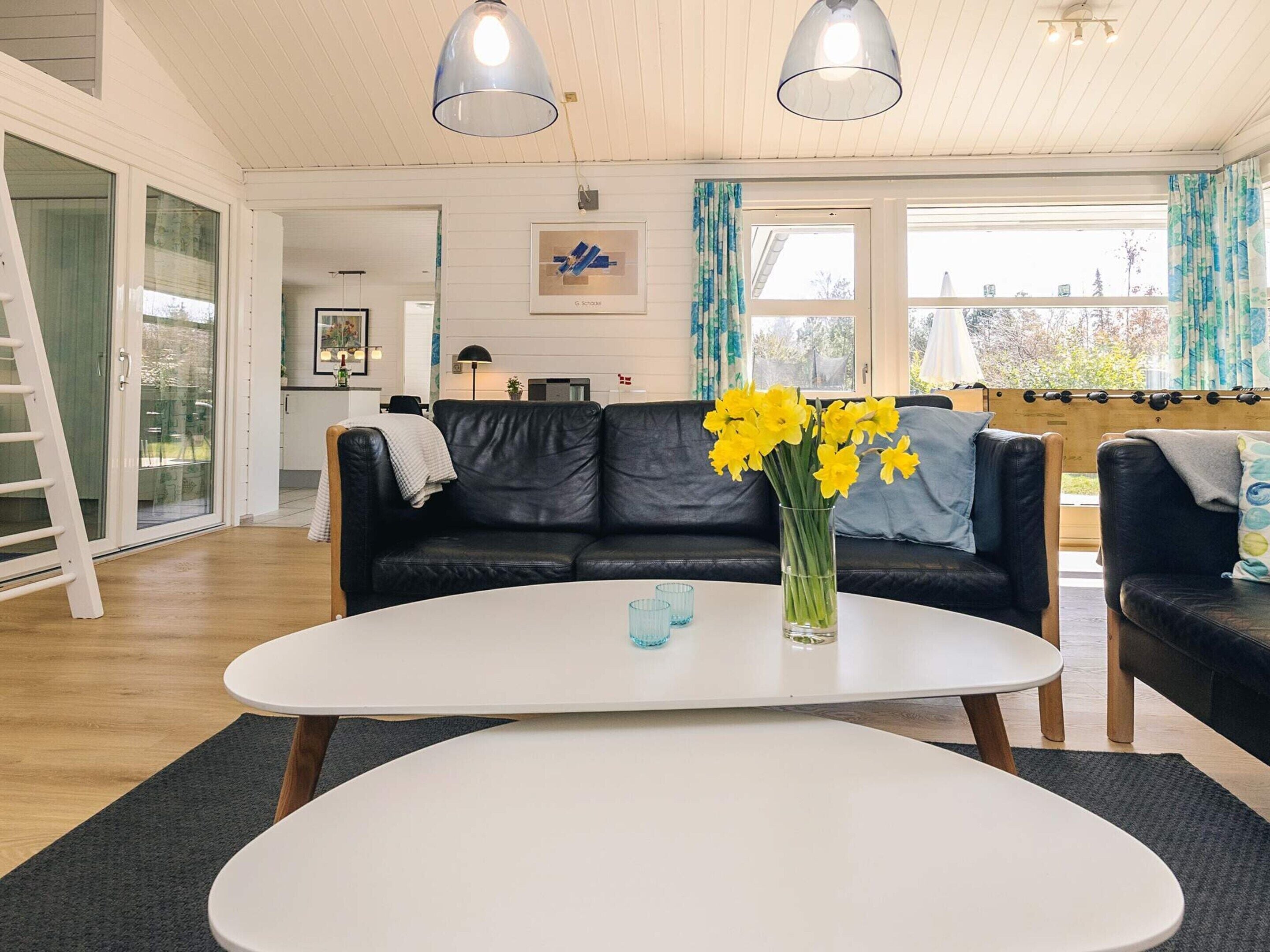Ett modernt vardagsrum med en svart lädersoffa, ett vitt soffbord och en vas med gula blommor. Ovanför bordet hänger två hänglampor, och det finns ett stort fönster med gardiner.