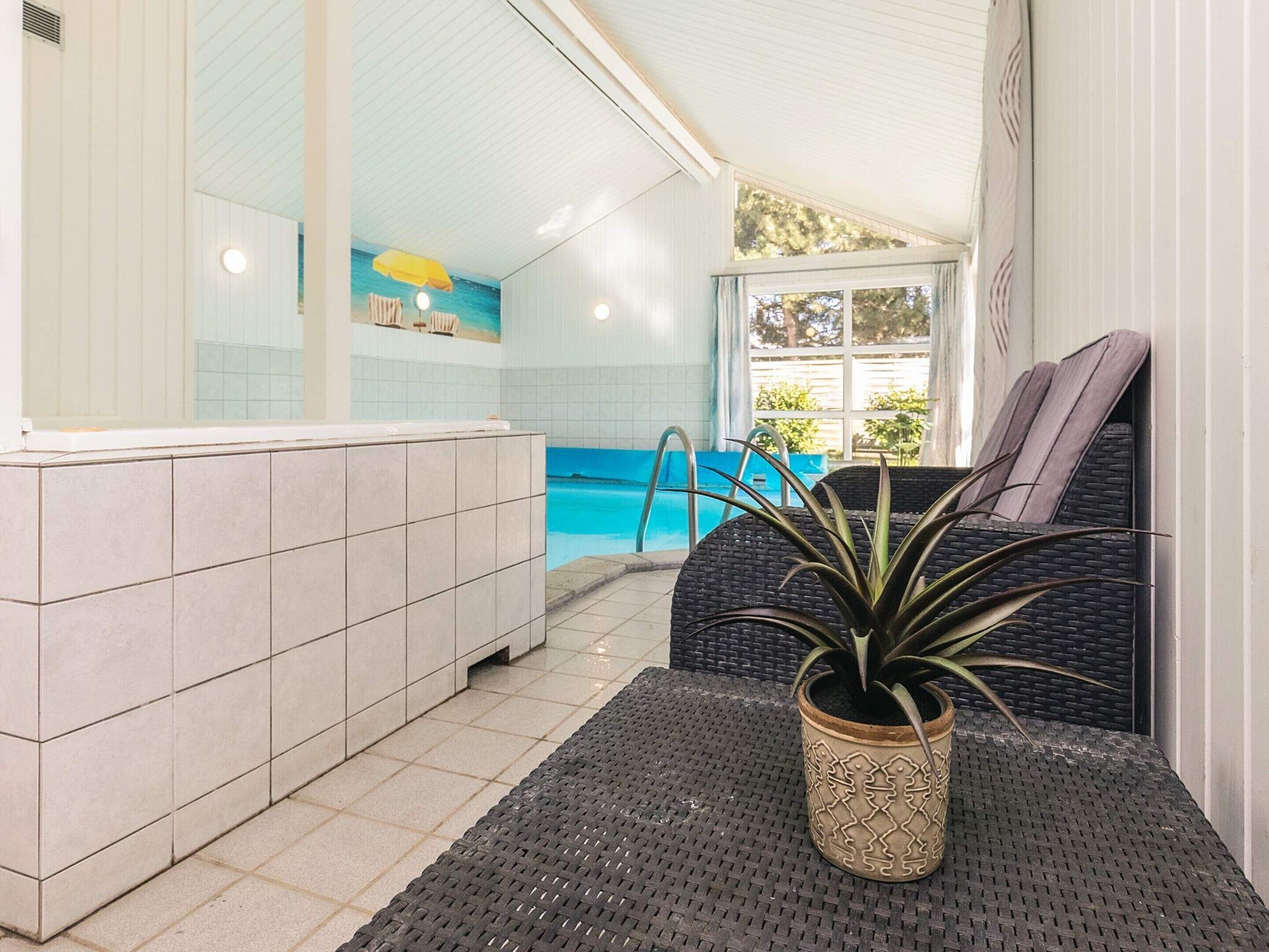 Un espace piscine doté d’un sol carrelé, d’une chaise longue, d’une plante en pot et offrant une vue sur la piscine à travers une fenêtre.