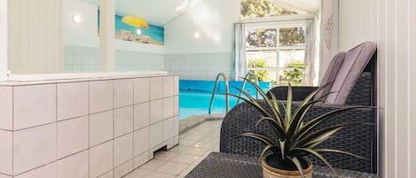 A pool area with a tiled floor, a lounge chair, a potted plant, and a view of the pool through a window.