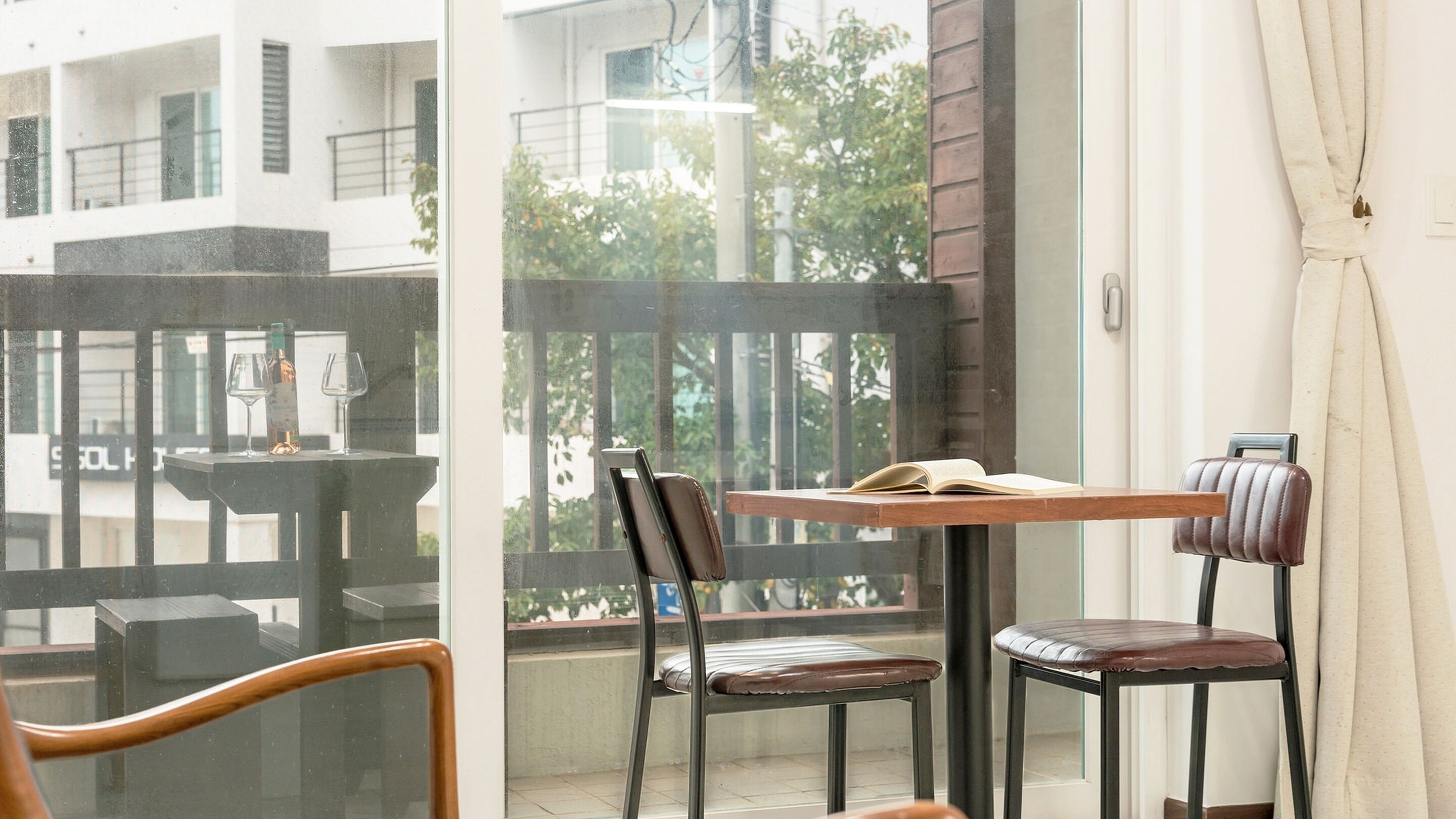 A balcony with a table and two chairs, a glass door, and a view of buildings and trees.