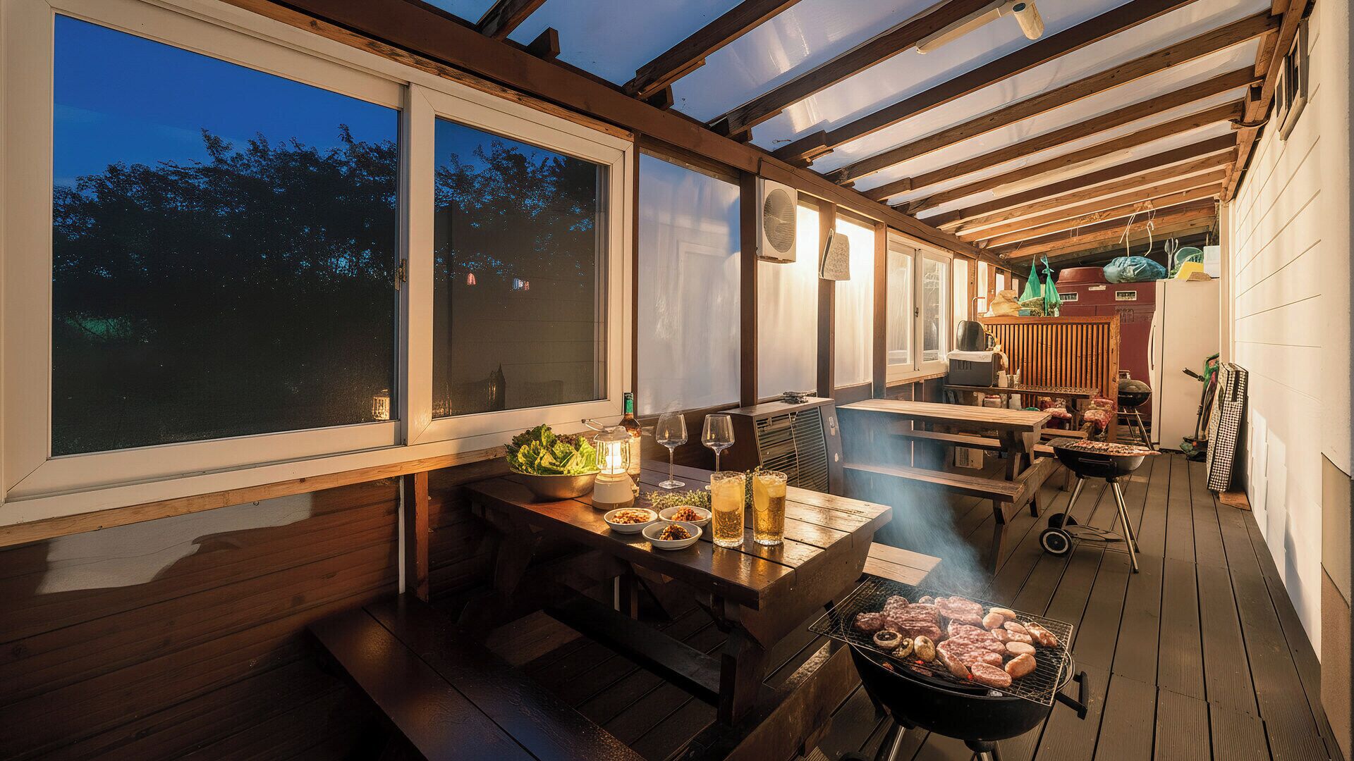 Un espace extérieur aménagé pour les repas, avec un barbecue, des tables en bois et une vue sur le ciel grâce à de grandes fenêtres.