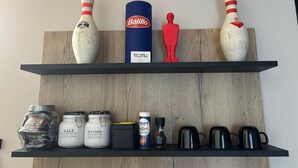 Ein Regal mit Bowlingkegeln, einer Balillo-Dose, einer roten Figur und Küchenutensilien wie Gläsern und Tassen.