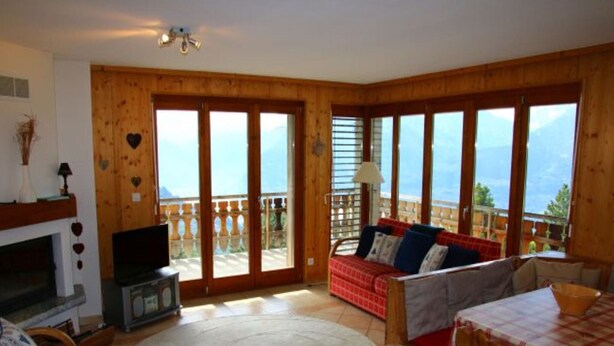Ein gemütliches Wohnzimmer mit Kamin, einer Couch, einem Esstisch und großen Fenstern mit Blick auf die Berge.
