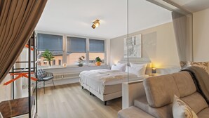 Habitación de hotel moderna con cama, sofá, escritorio y vistas a la ciudad a través de amplios ventanales.
