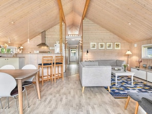 A spacious loft with a high ceiling, wooden beams, a kitchen with a dining area, a living room with a sofa and coffee table, and a hallway leading to another room.
