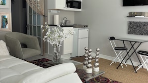 A modern living room with a white sofa, a colorful rug, a glass coffee table, and a kitchen area with white cabinets and a black microwave.