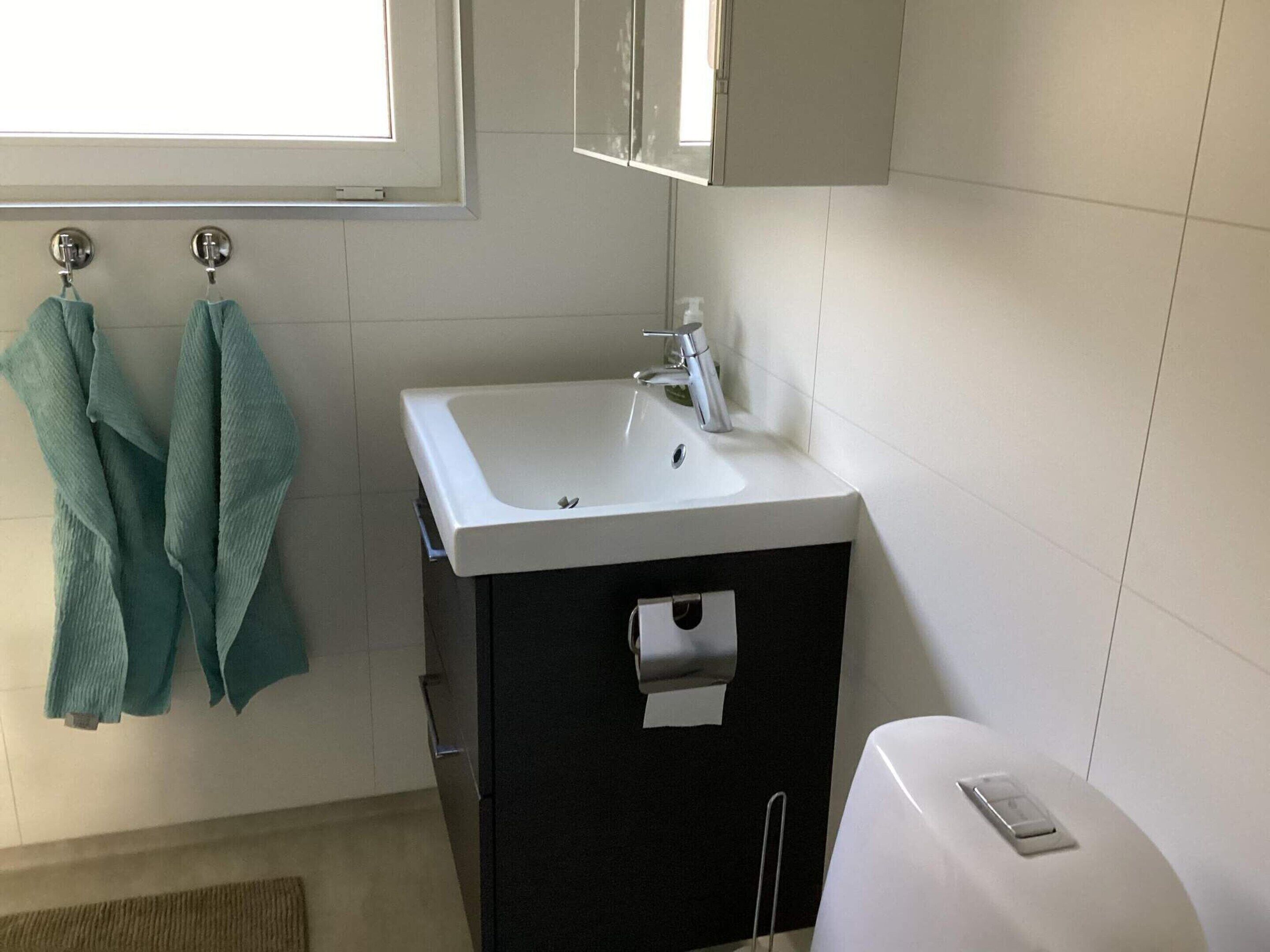 A bathroom with a white sink, a black cabinet, and a white toilet. There are two towels hanging on hooks.