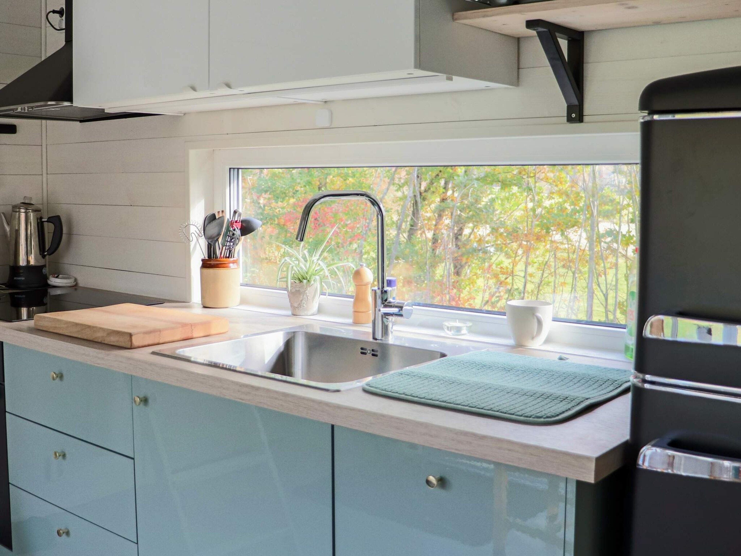 A modern kitchen with a stainless steel sink, a window with a view of trees, and a countertop with a cutting board and a plant.