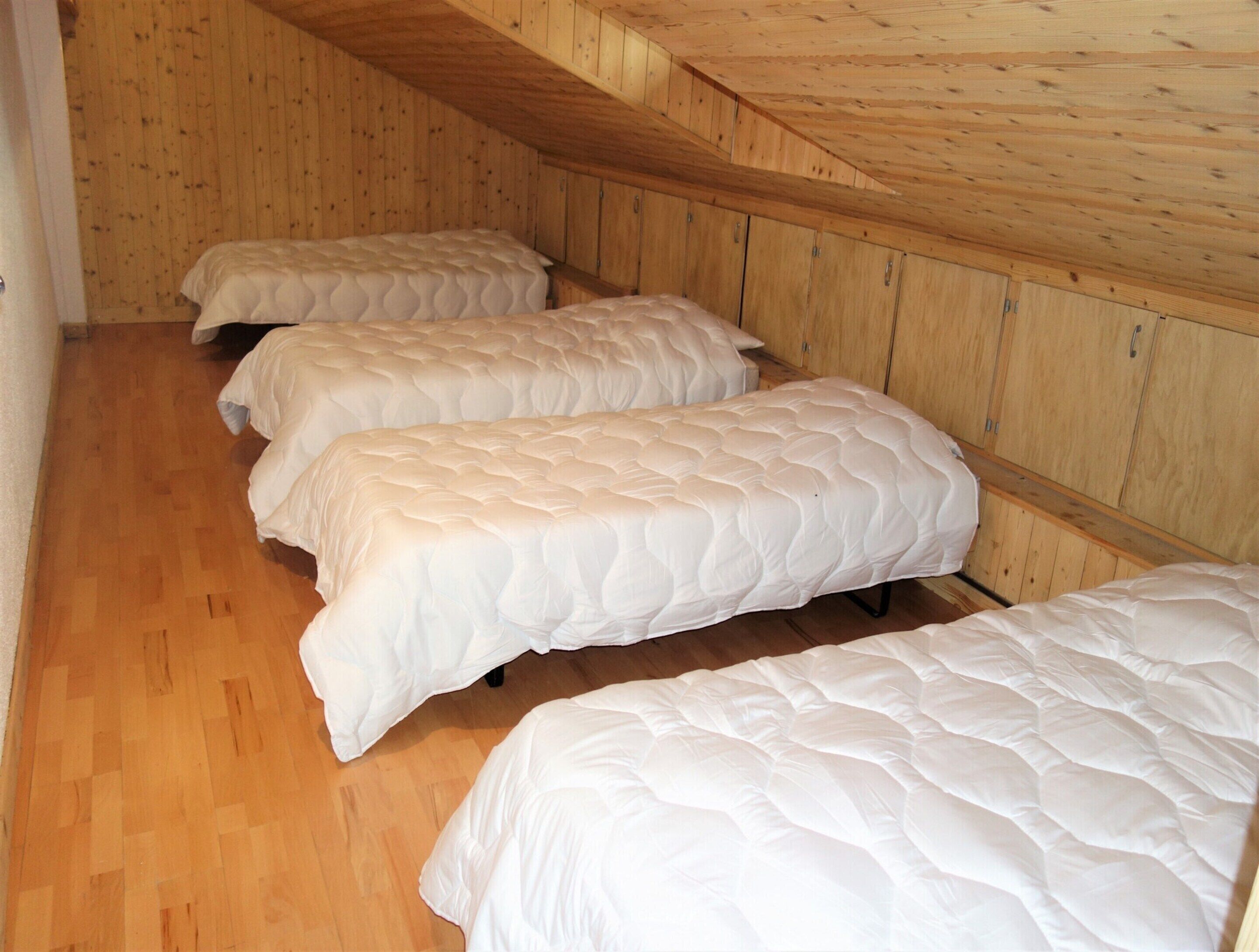 A room with three beds aligned against a wooden wall, wooden flooring, and a sloped ceiling.