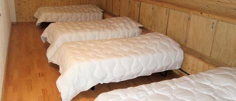 A room with three beds aligned against a wooden wall, wooden flooring, and a sloped ceiling.
