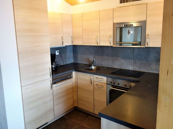 A compact kitchen with light wood cabinets, a black countertop, a built-in oven, and a stainless steel sink. - Ski Lift Apartment With Views (Veysonnaz)